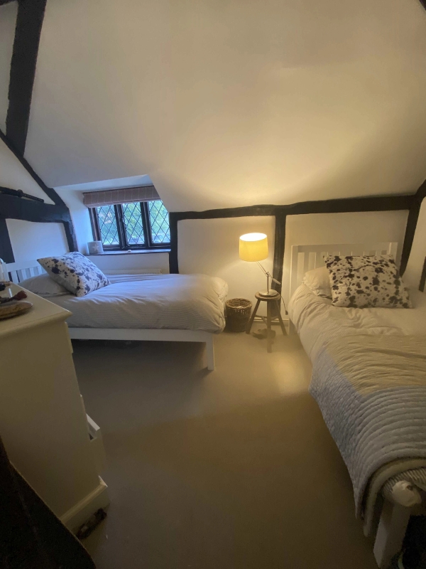Cozy twin bedroom space inside a historic Tudor cottage featuring warm bedside lighting and low-slung beamed ceilings, Love Locations.
