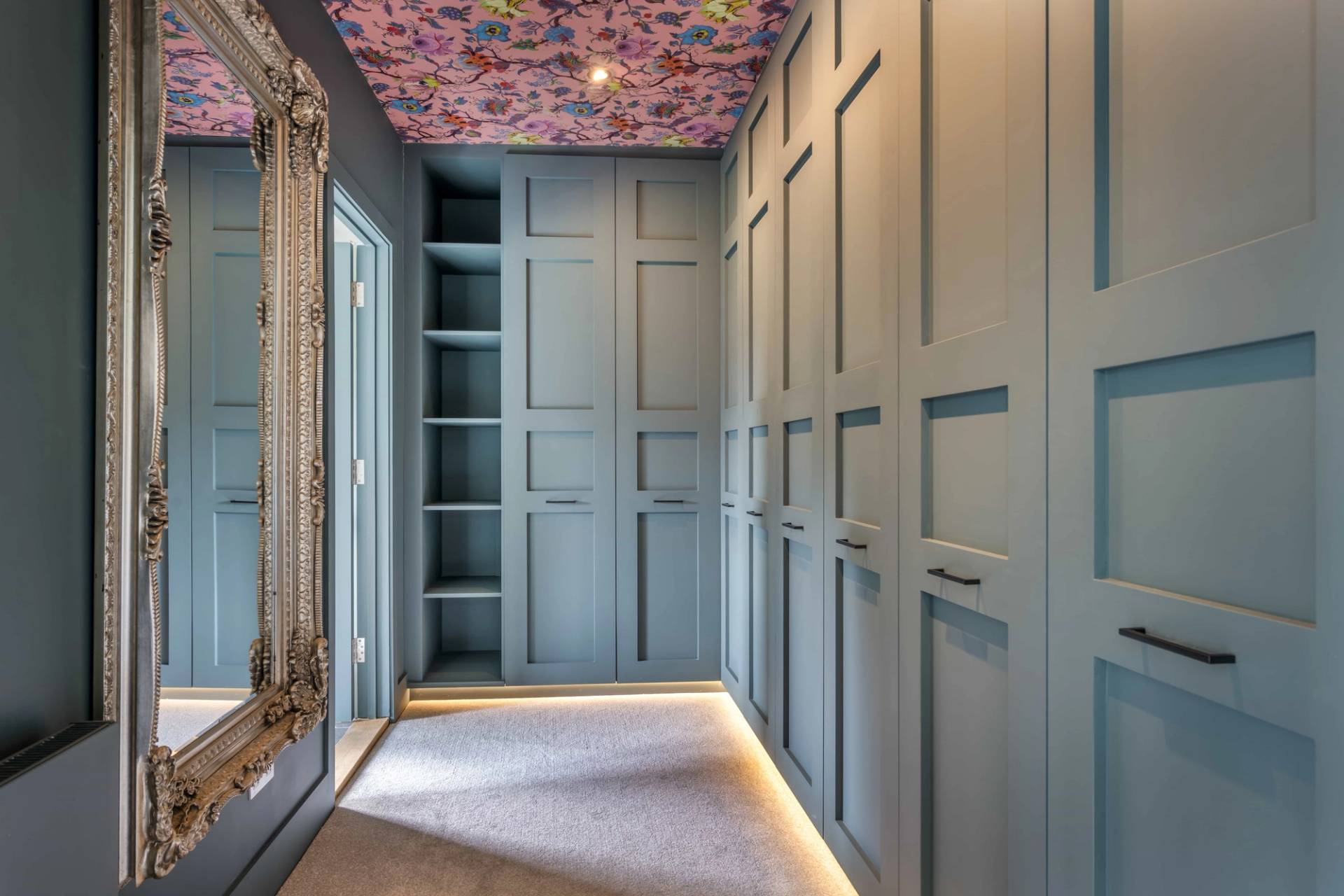 Bespoke blue dressing room closet featuring full-height built-in wardrobes, illuminated open shelves, and floral wallpaper ceilings, ideal for fashion brand shoots, Love Locations GU27.