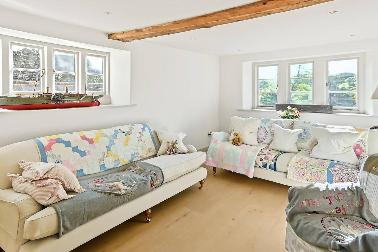 Quaint cottage sitting area location home in Gloucestershire GL7 from Love Locations, with low exposed timber beams, white plaster walls, daybed sofas, and colorful patchwork quilts.
