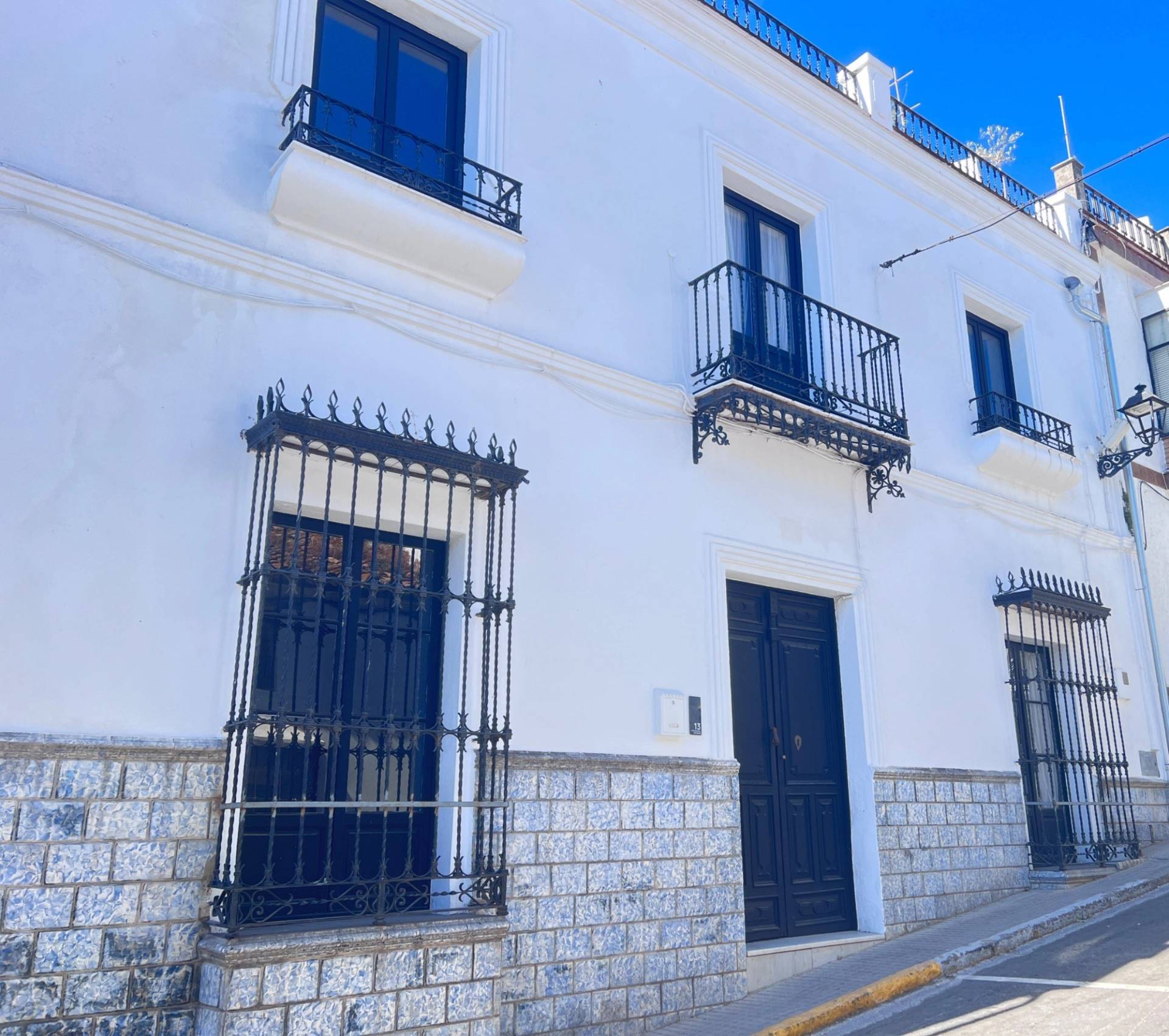 Authentic whitewashed Spanish townhouse exterior film location in Calle Sevilla Jimena de la Frontera Cádiz via Love Locations, showcasing traditional iron window grilles, dark entry door, and blue window frames.