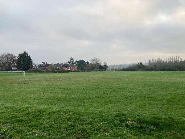 Love Locations large school playing field shoot space CM1: Sprawling green natural grass sports field under an open sky with a distant residential horizon backdrop, tailored for outdoor track and field filming.