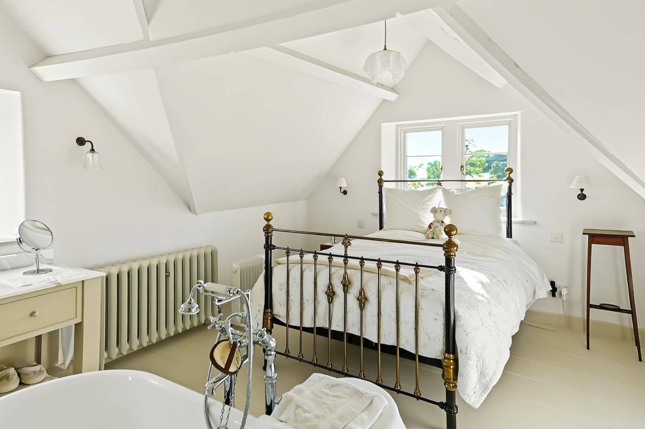 Airy attic bedroom film location in Gloucestershire GL7 through Love Locations, highlighting sloped vaulted ceilings, a wrought-iron double bedframe, and natural sunlight.