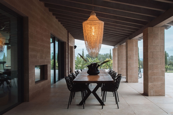 Covered outdoor patio terrace with long dark wooden dining table, black chairs, and woven pendant light fixture, Love Locations 07630.