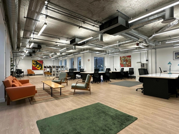 Bright open office space with industrial-style exposed ceiling and metal staircase, featuring a bright orange sofa, modern chairs, small coffee table, and green rug in the foreground.