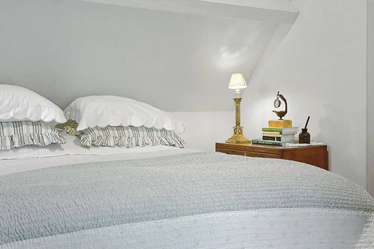 Cozy bedside nook photoshoot location in Gloucestershire GL7 by Love Locations, featuring soft white linen bedding, a wooden nightstand, and a warm reading lamp under sloped eaves.