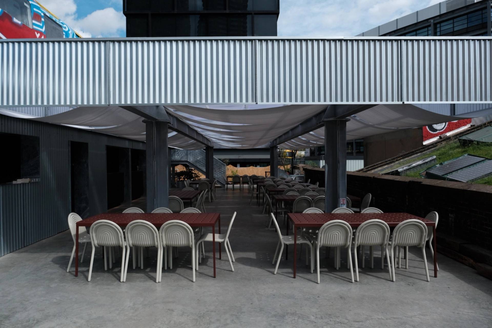 Covered outdoor courtyard seating area with steel pillars, long wooden tables, and white chairs via Love Locations.