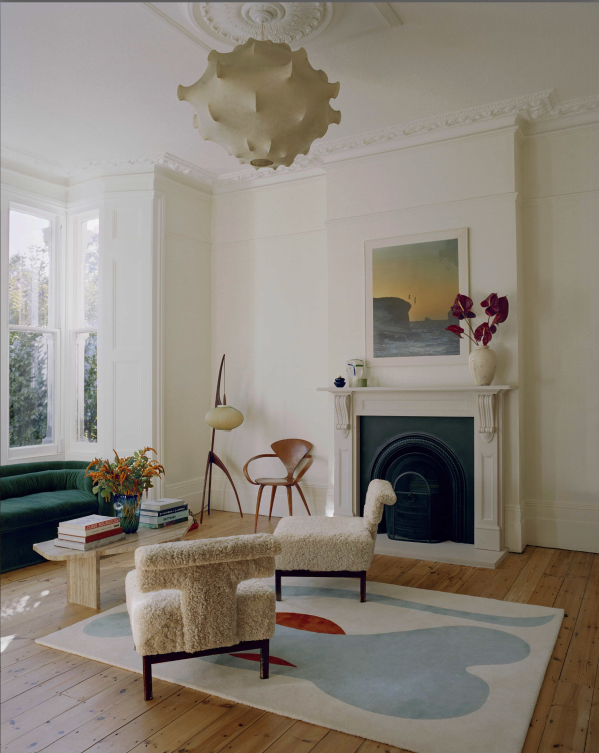 A bright period living room with high ceilings, decorative cornicing, a white fireplace, sculptural boucle lounge chairs, a curved green velvet sofa, abstract rug, and oversized statement pendant light.