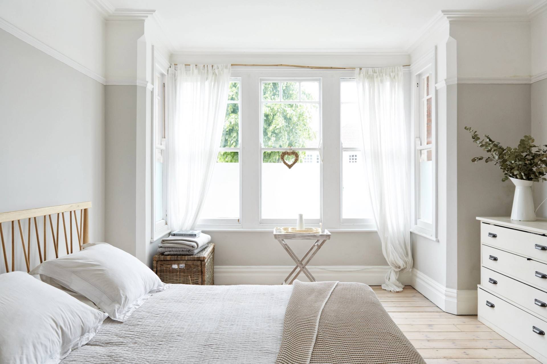 Light-filled bedroom corner featuring white sheer curtains, neutral bedspread, and bedside storage table, Love Locations N13.