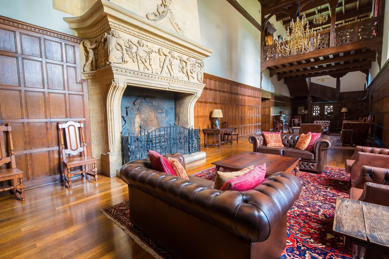 Dramatic Jacobean grand hall living room location in Hertfordshire AL9 from Love Locations, featuring a deep brown leather Chesterfield sofa, monumental stone fireplace, and high timber-beamed ceiling.