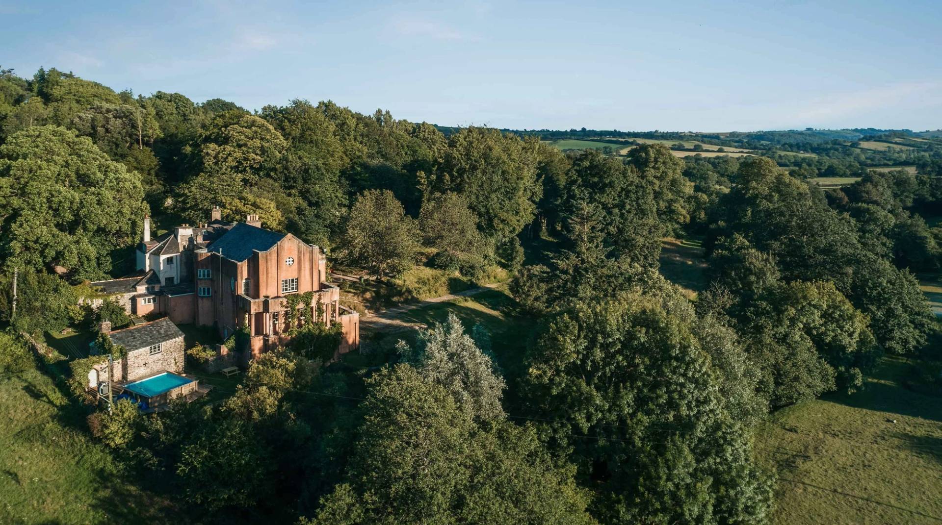Elevated aerial view of a red brick country property nestled among dense green woodland in Totnes Devon TQ12 through Love Locations.
