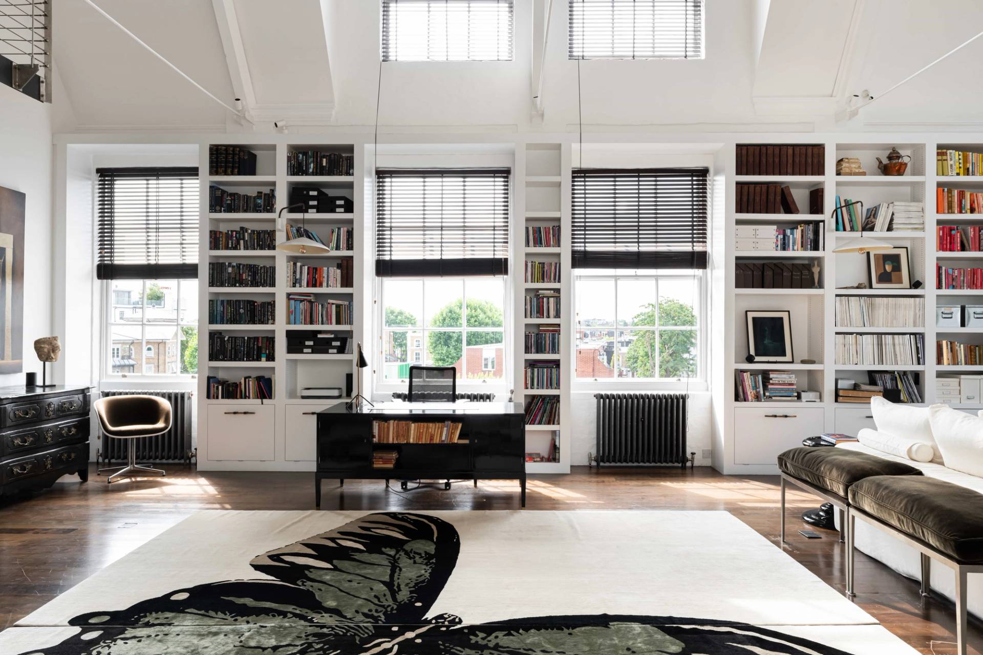 Home office study area in a vaulted ceiling loft featuring a dark wood desk, black radiator, and full-height white bookshelves, Love Locations SW11.