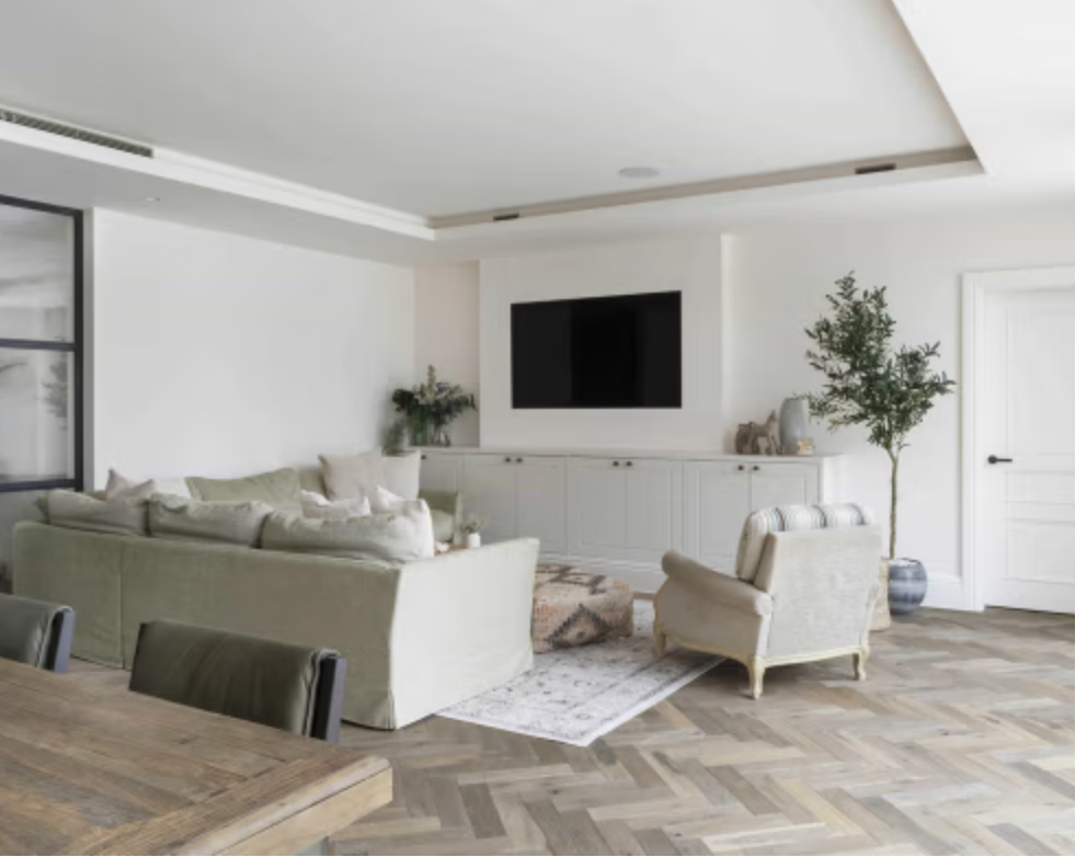 Modern family living room at Badgers Drift with a large neutral sofa, herringbone wood flooring, bespoke built-in cabinetry, wall-mounted television, and a bright open-plan layout.