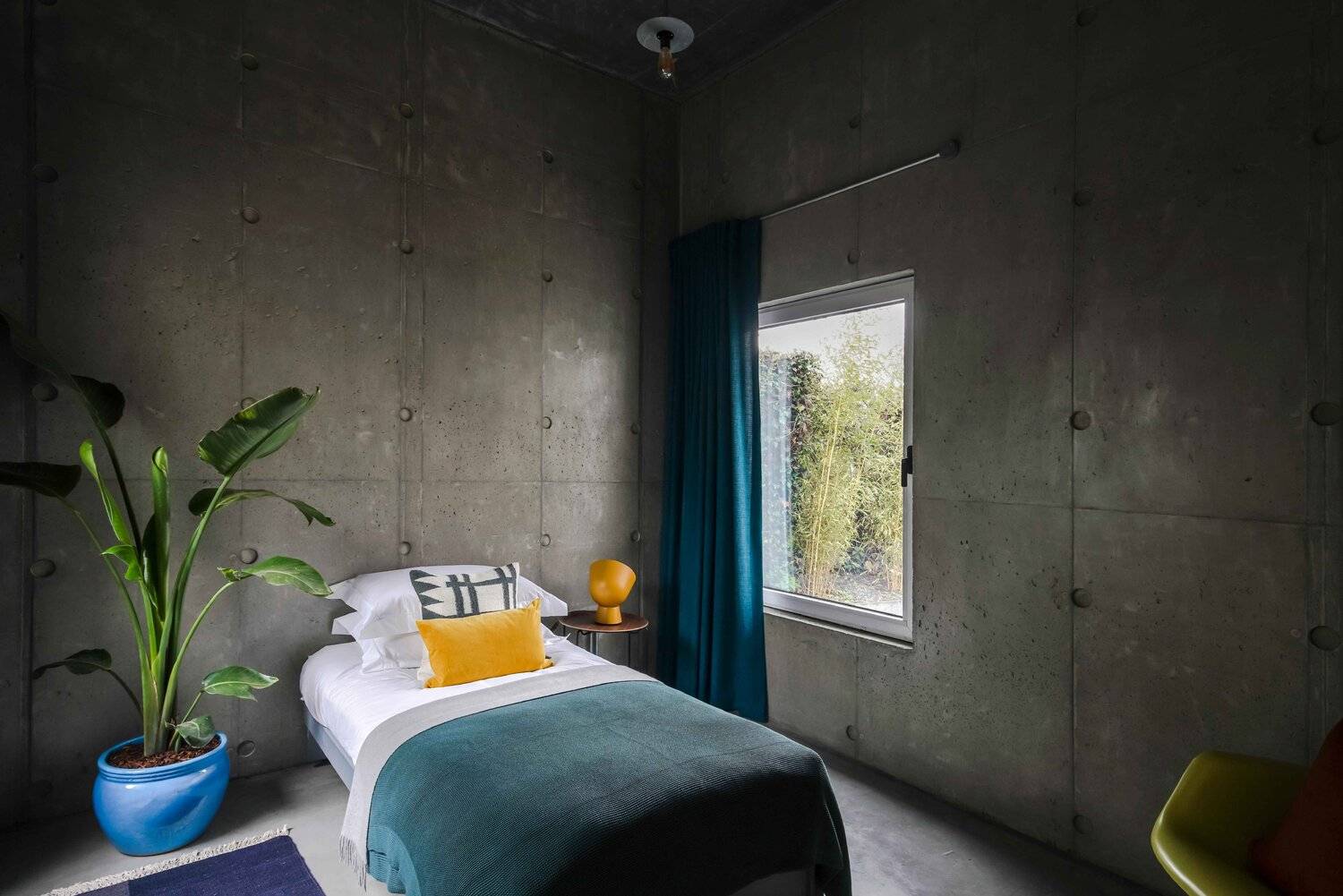 Minimalist bedroom featuring raw concrete walls, single bed with teal throw, yellow lamp, and window light, Love Locations BN7.