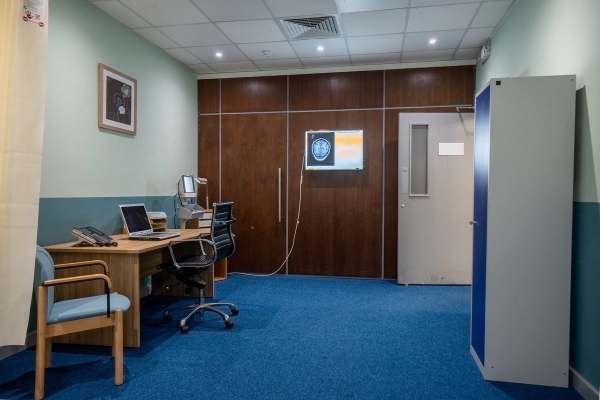 Doctor consulting room with office desk, computer monitor, blue carpet, and wood-panelled accent wall, Love Locations UB4.