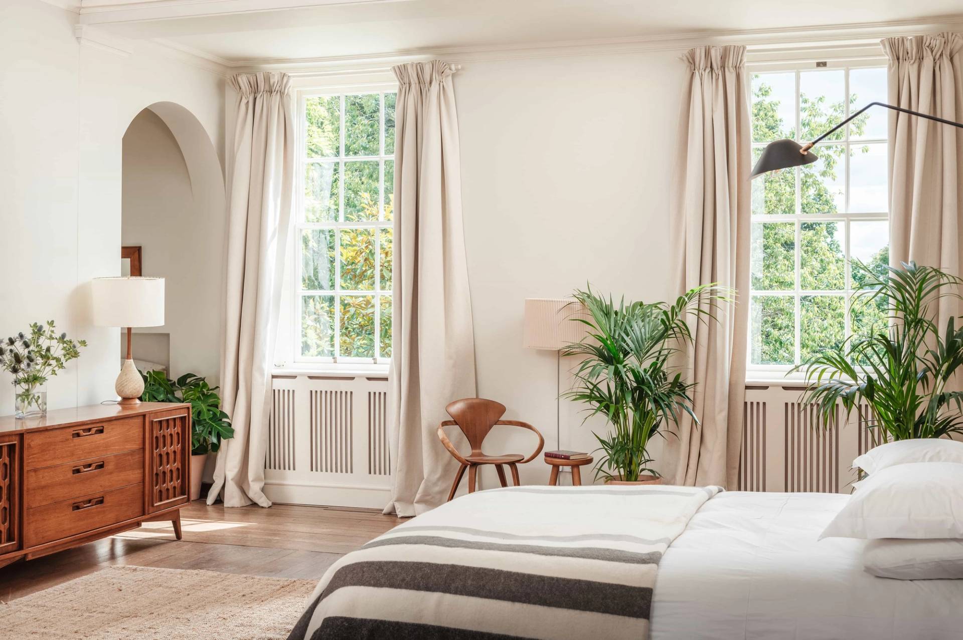 Sunlit bedroom corner with full-length drapes, light parquet flooring, and indoor greenery in a Devon location home from Love Locations.