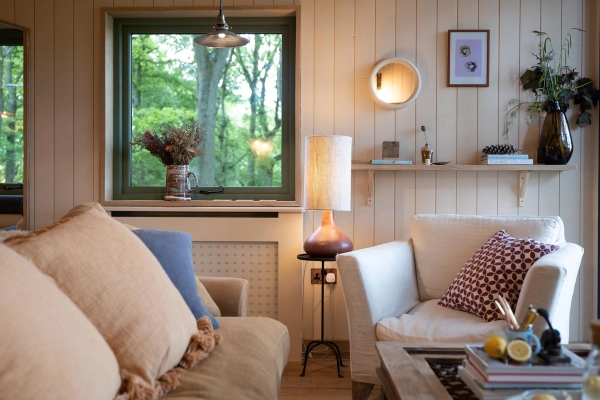 Comfortable seating corner inside a luxury cabin featuring a tan sofa, white armchair, and soft lamp lighting, bookable through Love Locations Herefordshire.