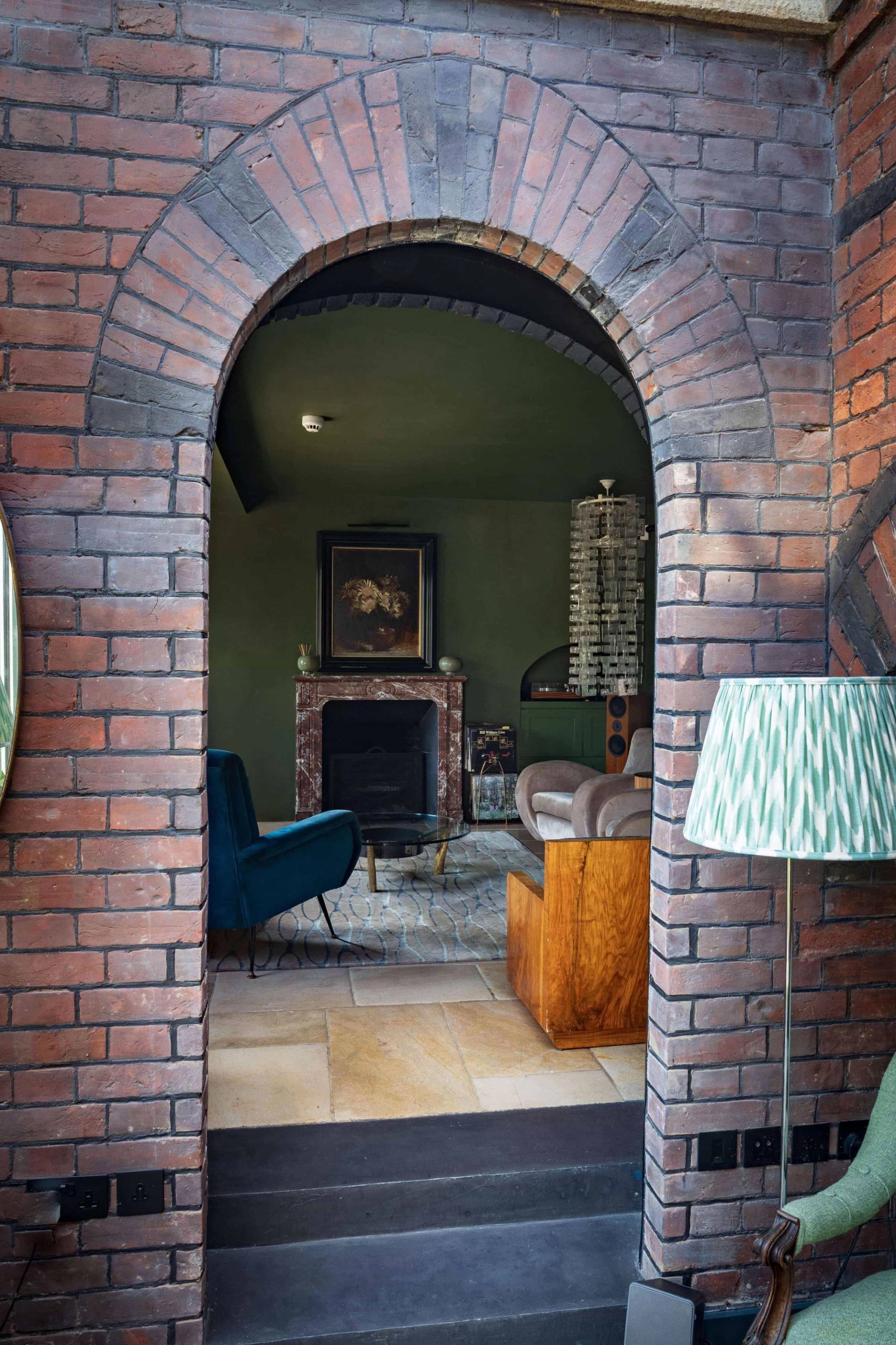 View through a red brick arch into an atmospheric living room with a marble fireplace and blue armchairs, Love Locations.