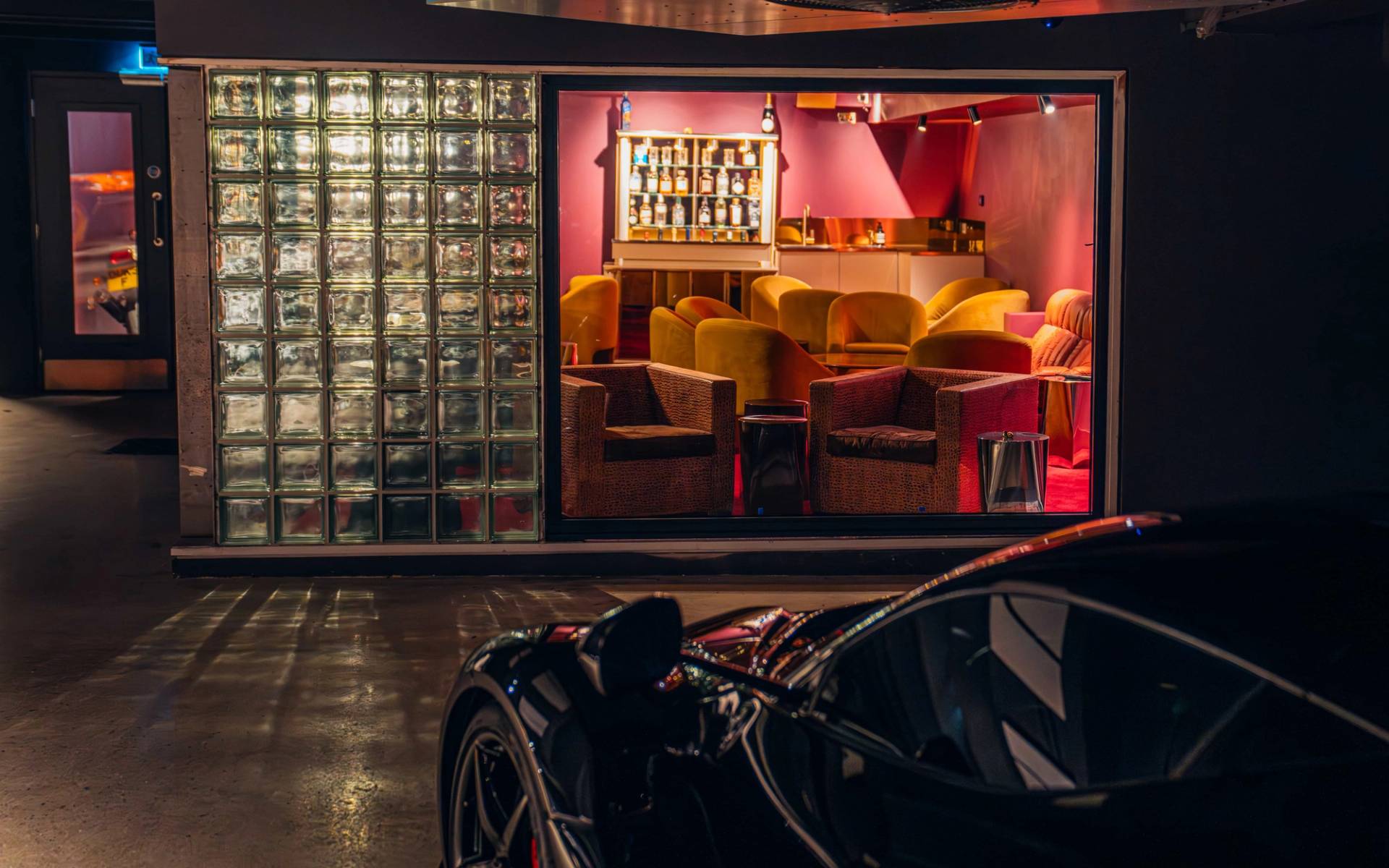 Glass block partition wall framing a warm red velvet cocktail lounge view with a black hypercar in the foreground by Love Locations.