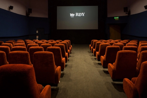 Symmetrical view down the carpeted aisle towards the illuminated projection screen displaying the Ritzy logo inside The Ritzy Screen 3 film location in Brixton SW2 from Love Locations, featuring plush red velvet seats.