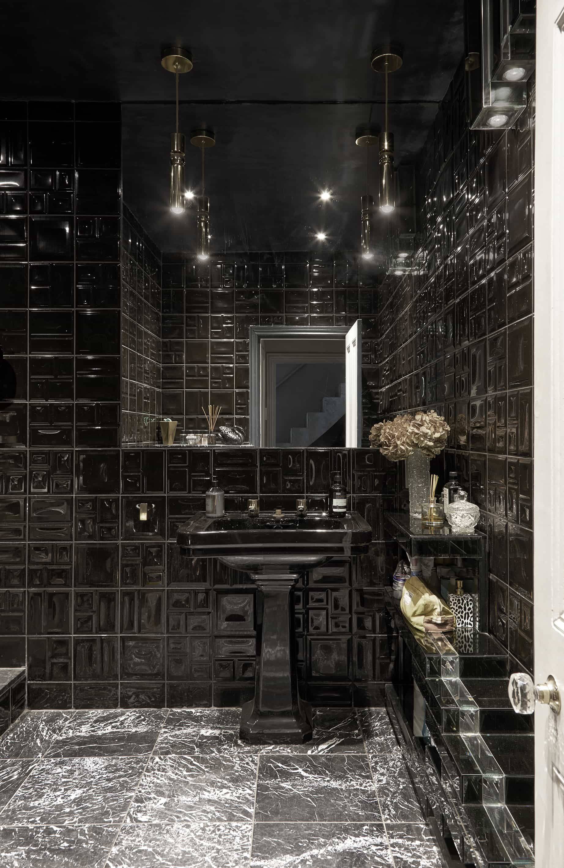 Dramatic dark moody bathroom featuring glossy black square wall tiles, black pedestal sink, and silver mirror, Love Locations N16.