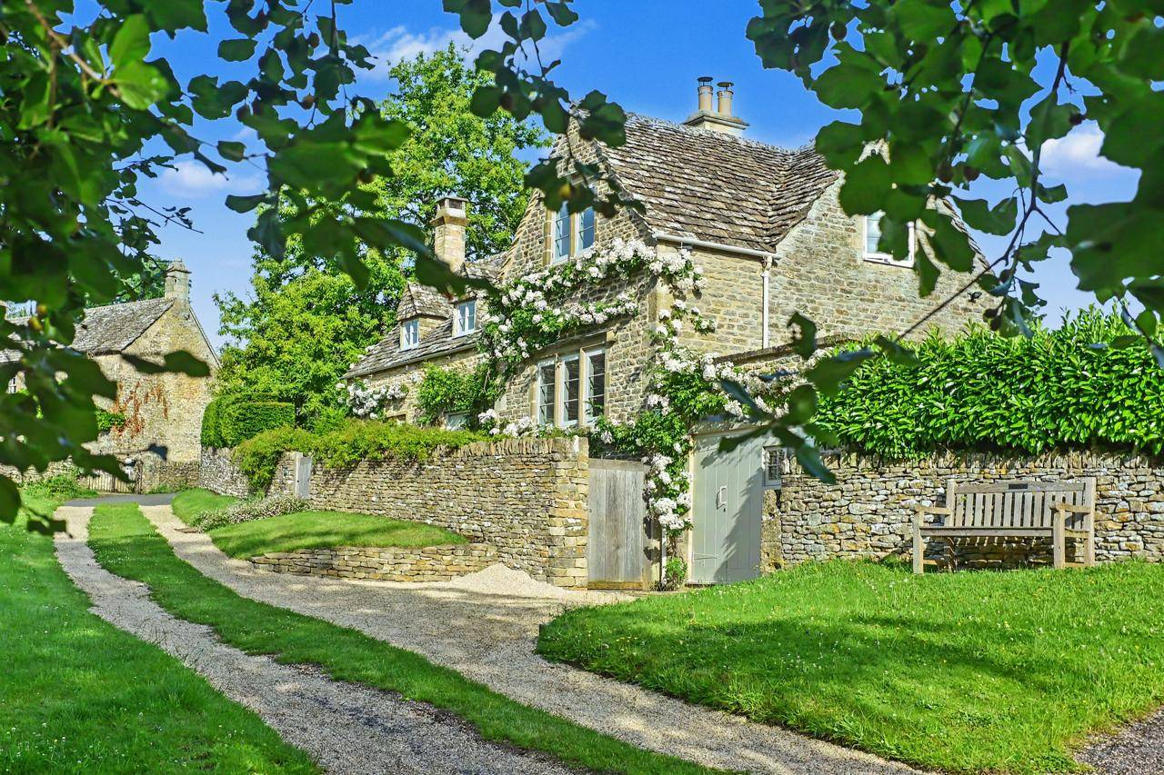 Exterior view of a Cotswold stone cottage with climbing ivy film location in Gloucestershire GL7 via Love Locations, featuring a gravel pathway, stone walls, and lush green foliage.