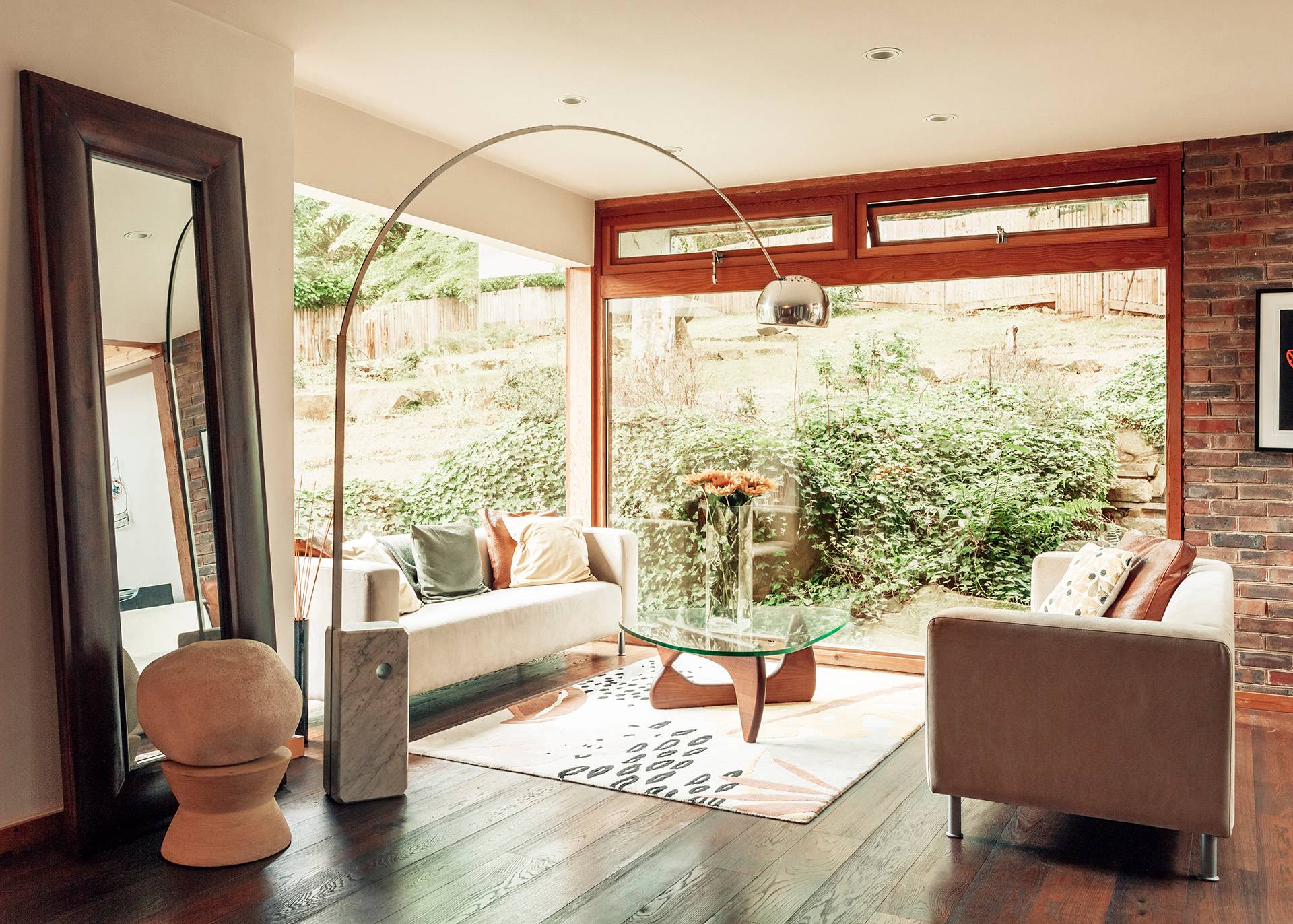 Cozy mid-century garden sitting room featuring large windows, a neutral sofa, and an arc floor lamp overlooking greenery, Love Locations.