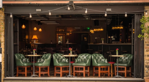 Line of luxury tufted green leather armchairs arranged along an outdoor-facing terrace counter under festoon lighting, suitable for high-end casual dining shoots, Love Locations.