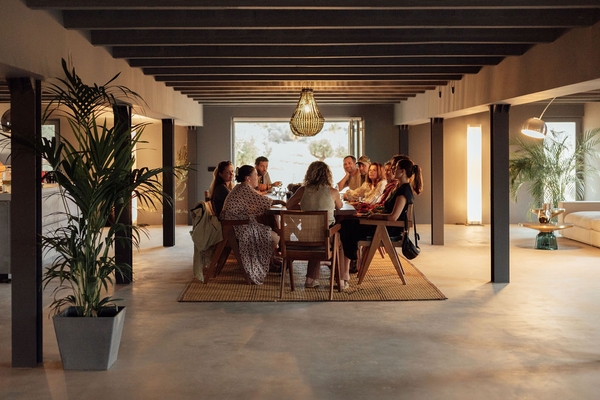 Open-plan villa reception room with people seated around a long wooden dining table, Love Locations 07630.