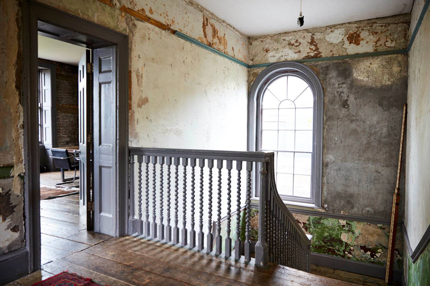 Spacious stairwell landing with dark blue banisters, arched sash window, and distressed plaster walls, Love Locations.