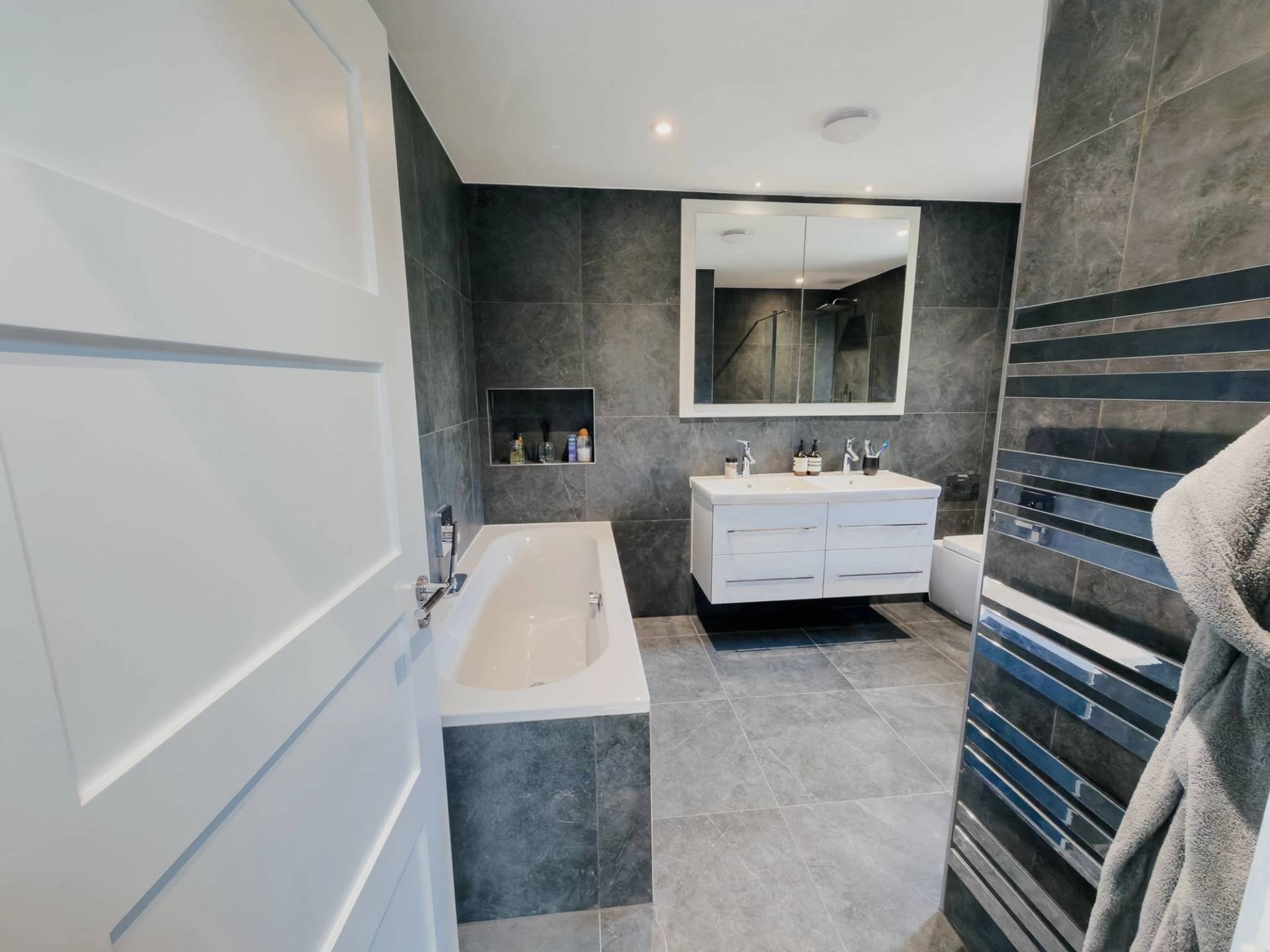Modern grey marble bathroom shoot space in Barnet EN4 via Love Locations, featuring a built-in bath, white double vanity unit, and dark slate wall tiling.