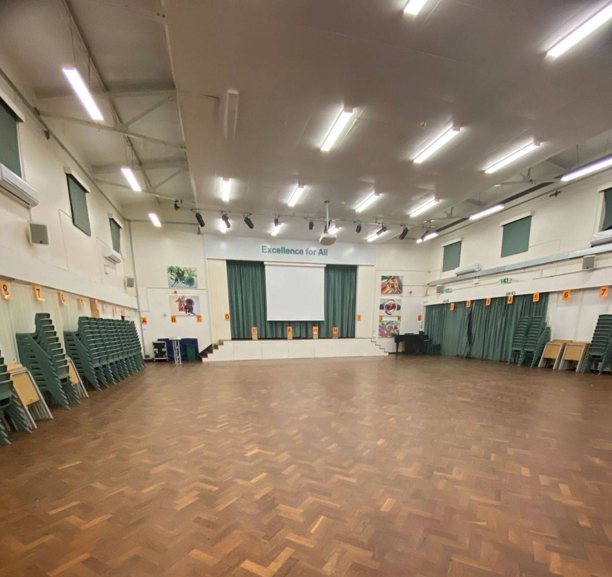 Wide view of an empty school hall photo location displaying parquet flooring and stacked green chairs along the walls by Love Locations.