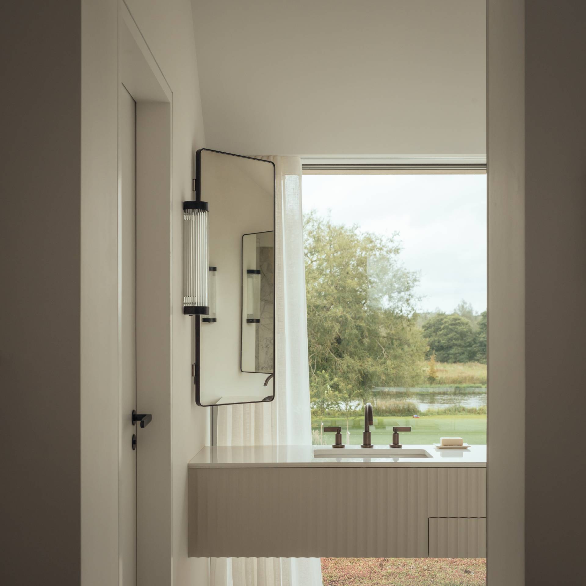 Luxury bathroom mirror vanity setup framed by custom lighting and large picture windows looking onto greenery, Love Locations.