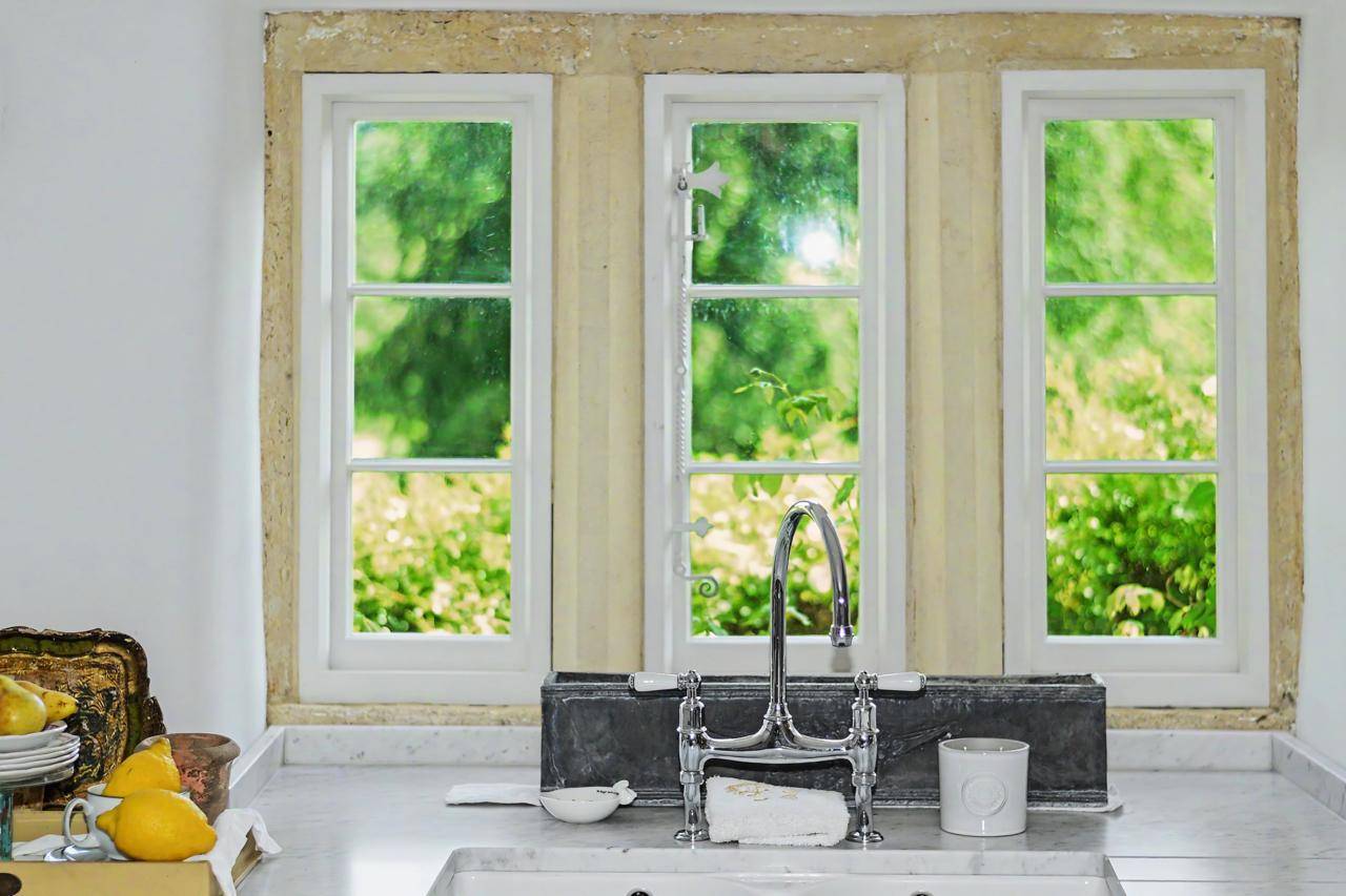 Cottage window sill and sink view film location in Gloucestershire GL7 via Love Locations, looking out onto bright green garden foliage through white multi-pane windows.