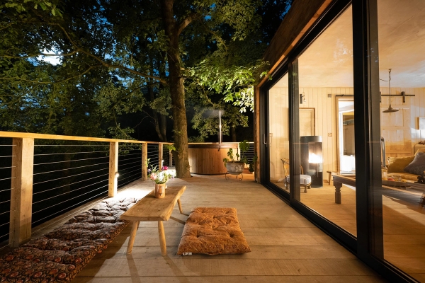 Atmospheric twilight view of an illuminated timber deck veranda with floor cushions next to glowing cabin windows, Love Locations Herefordshire.