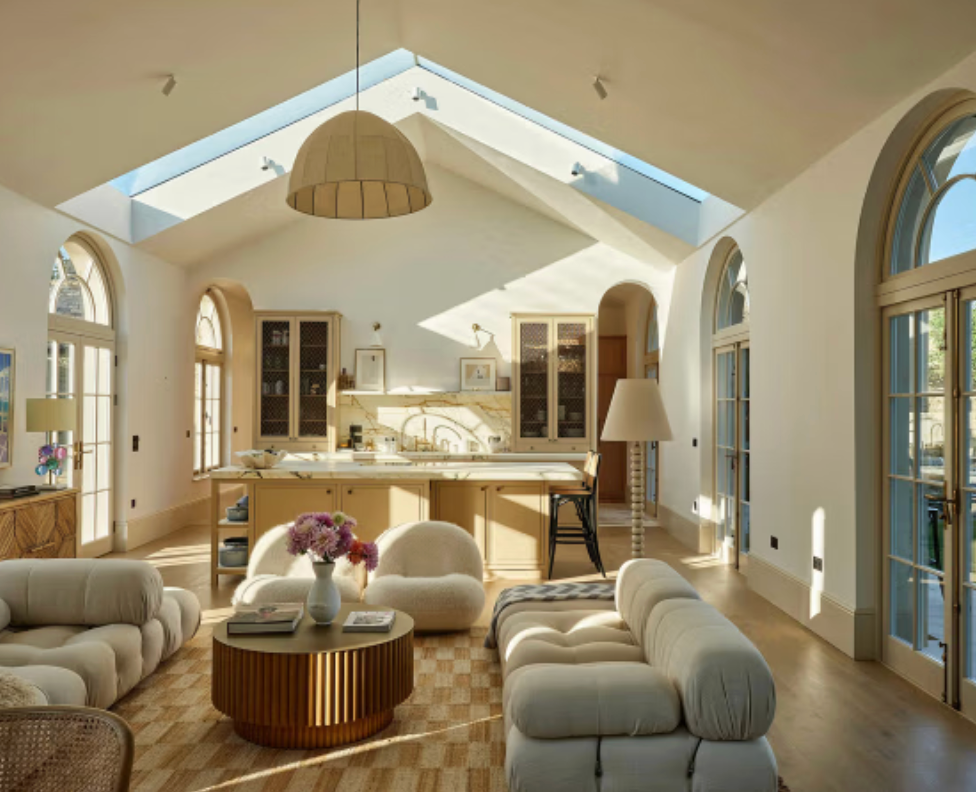 Open-plan kitchen and living area at Cotswold Farm featuring vaulted ceilings with skylights, arched steel-framed doors, a contemporary kitchen, curved boucle sofas, and warm natural finishes.
