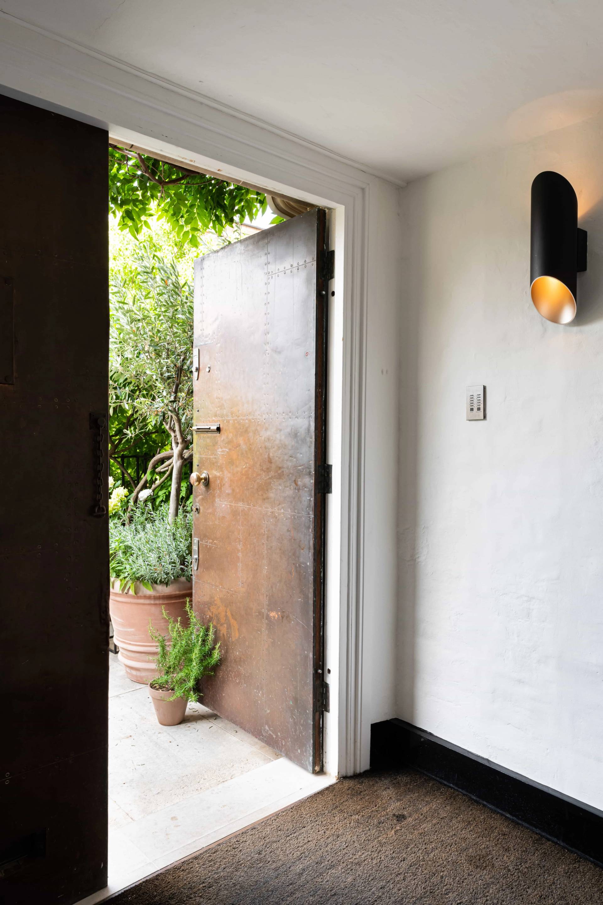 Heavy industrial metal entrance door opening into a garden courtyard with black wall sconce lighting, Love Locations.