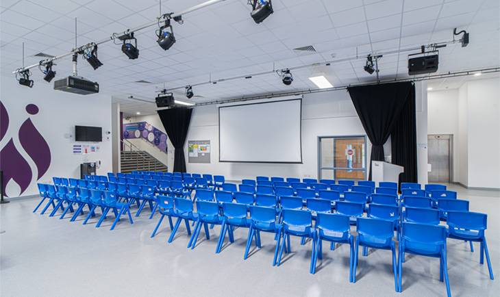 Large school assembly hall film location in Hove BN3 by Love Locations, featuring rows of blue plastic chairs, stage lighting rig, and projection screen.