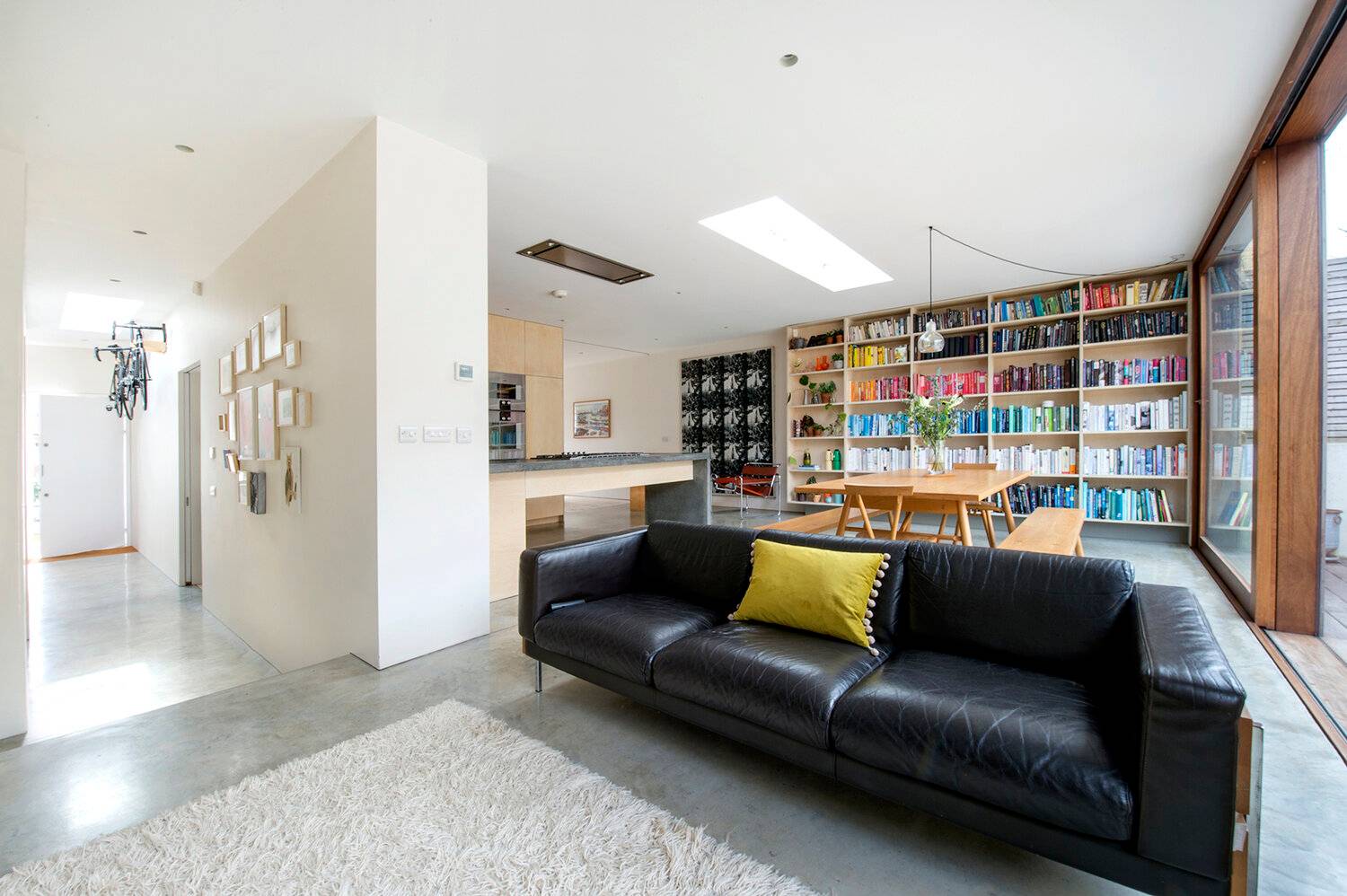 Spacious open-plan living room with a black leather sofa, polished concrete floors, and full-wall plywood bookcase, Love Locations SE23.