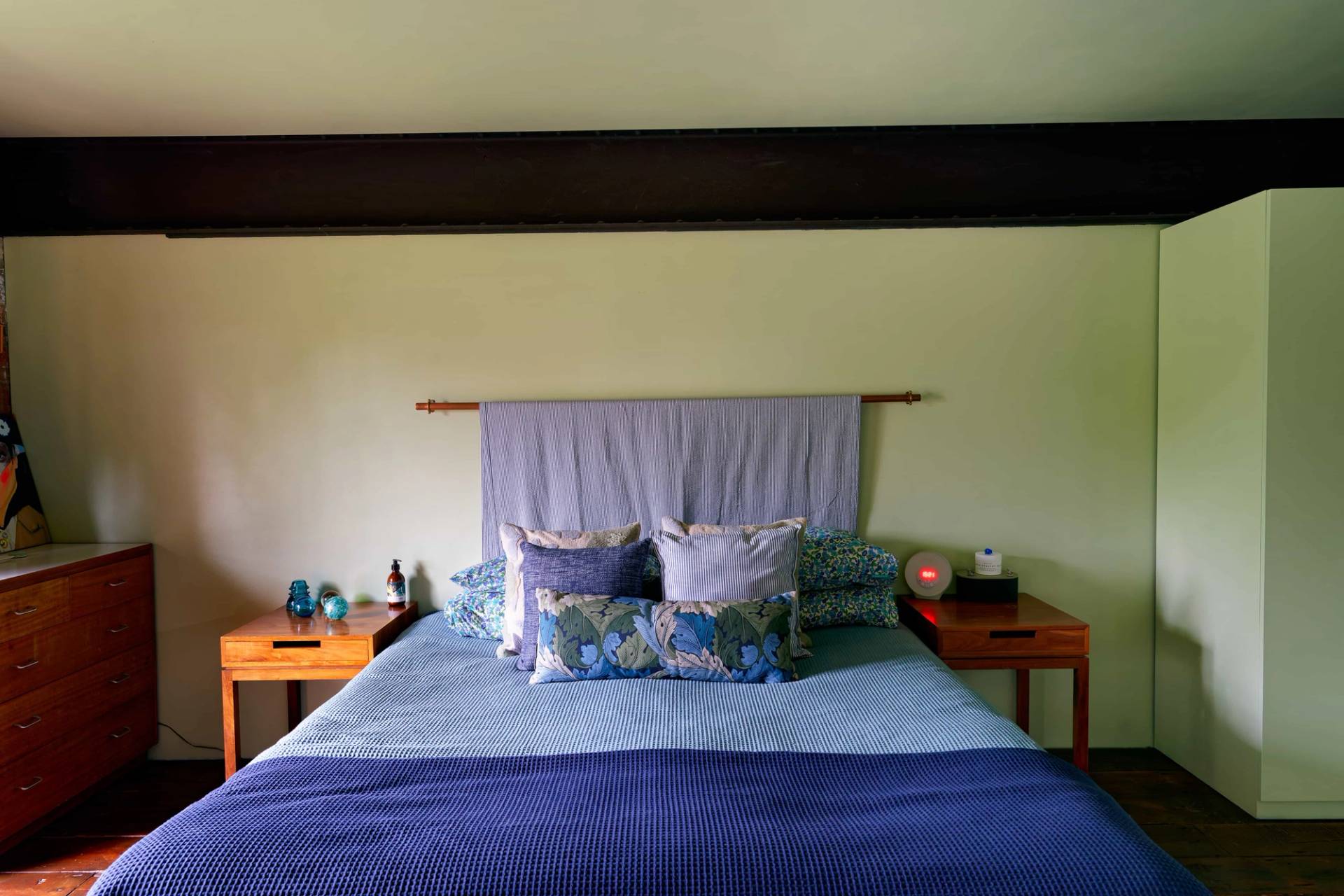 Loft bedroom setup featuring dark timber floors, sage green walls, and blue linen bedding set under exposed ceiling beams, Love Locations.