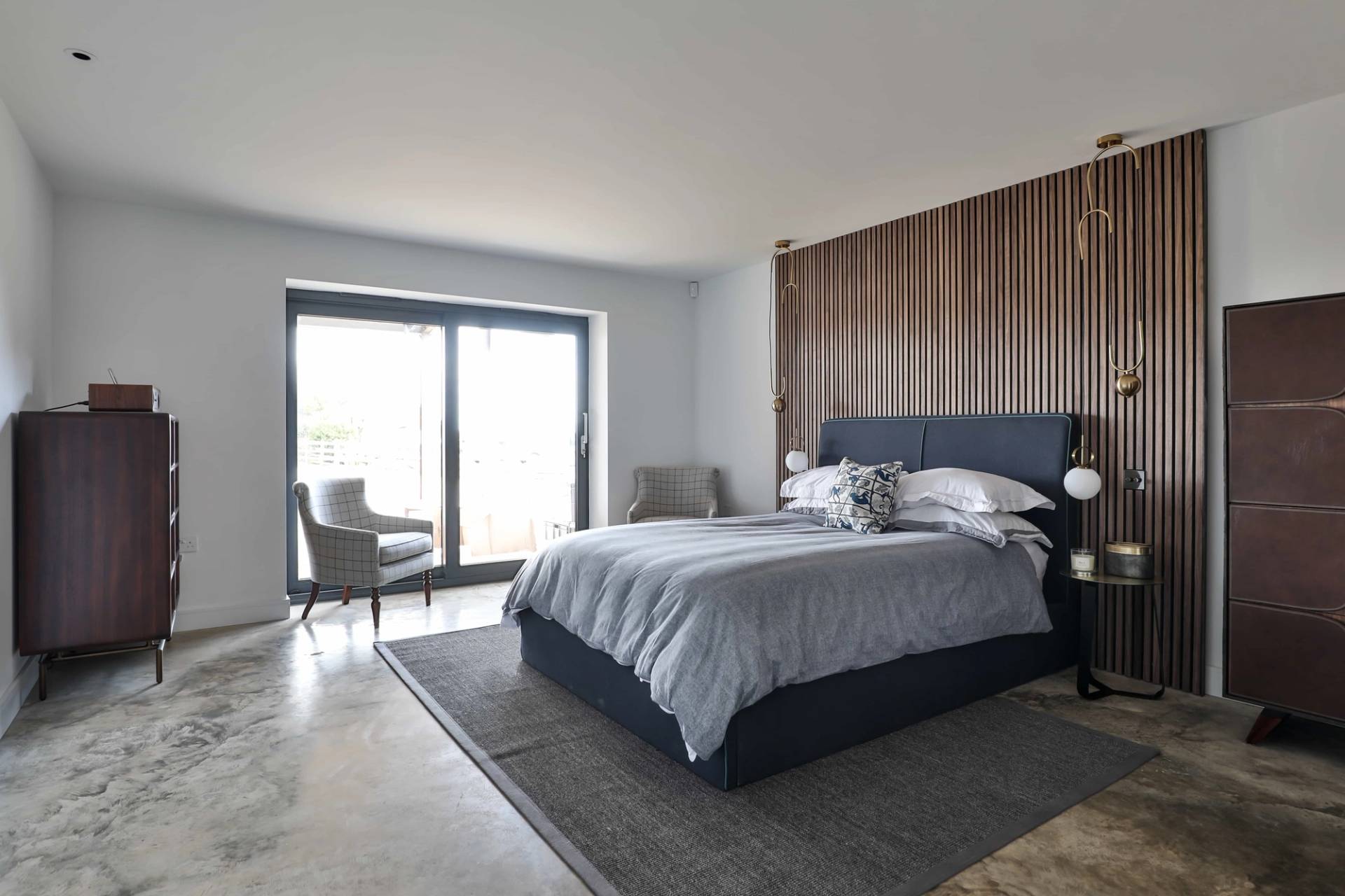 Sunlit bedroom suite with slatted timber headboard wall, area rug, and sliding patio doors, Love Locations CV23.