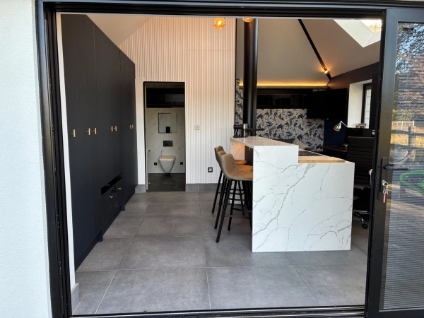 Contemporary pool house kitchenette and bar location in Essex CM4 via Love Locations, featuring sleek black cabinetry, a white marble kitchen island, and light tiled floors.