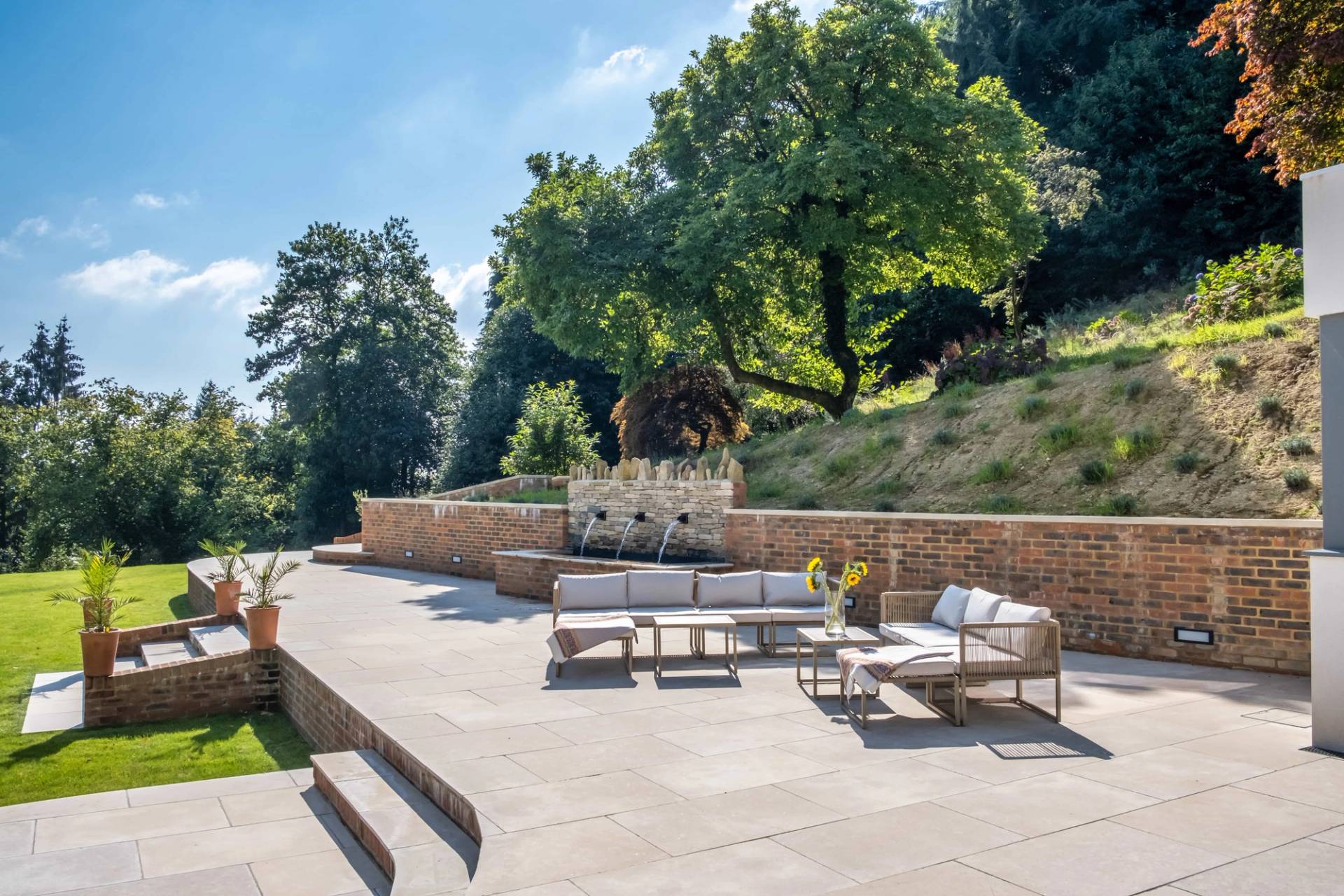 Stone paved garden terrace with outdoor lounge seating set against brick retaining walls and woodland grounds, Love Locations.
