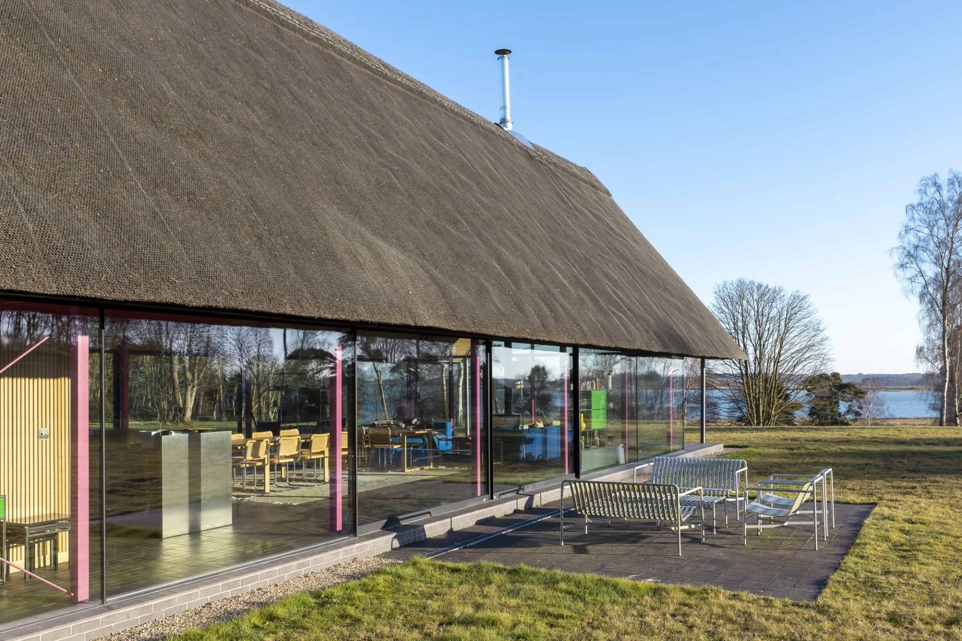 Exterior view of a modern thatched longhouse featuring full-height glass walls set against manicured green lawns, Love Locations IP17.