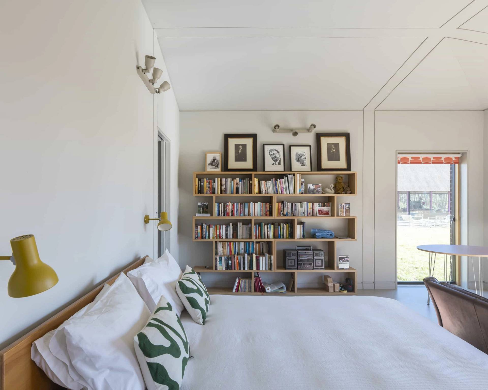 Bright white master bedroom featuring a wooden bed frame, green palm leaf cushions, and built-in bookshelf storage, Love Locations IP17.