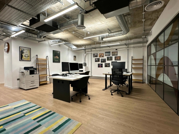 Bright open office space with industrial-style exposed ceiling,glass feature wall, desk spaces, large notice board, and a striped rug