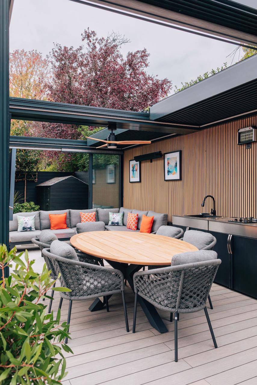 Modern outdoor lounge pergola with grey corner sofa, orange accent cushions, and built-in outdoor kitchen, perfect for summer campaigns, Love Locations W5.