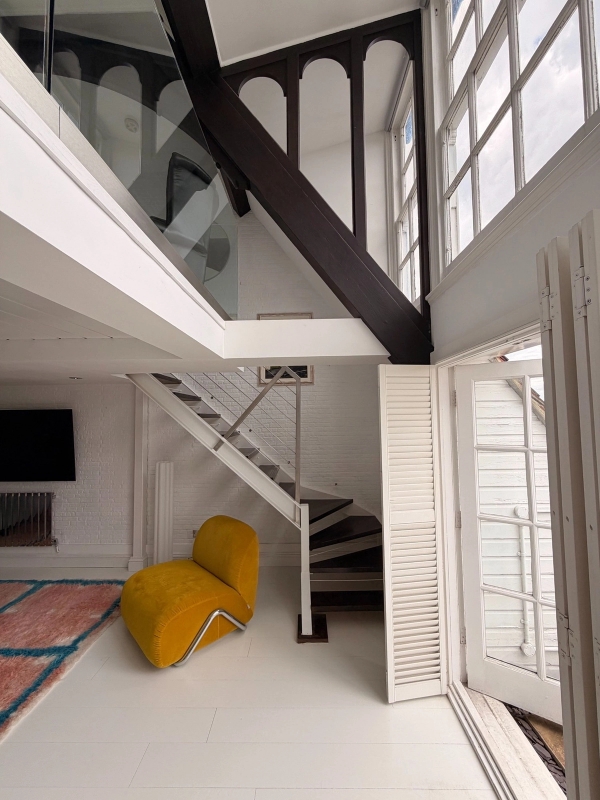 Double-height loft living room featuring exposed dark steel beams, a white floating metal staircase, and a mustard yellow statement chair, curated by Love Locations.