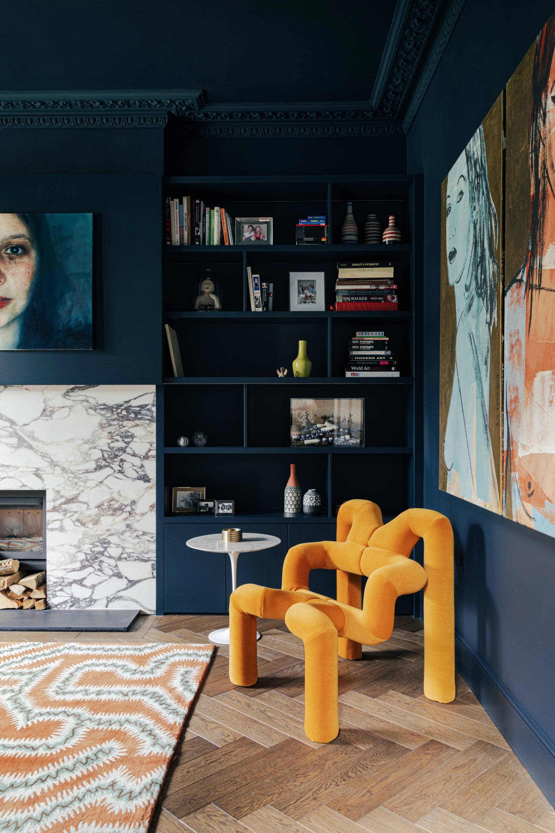 Moody blue reception room featuring a marble fireplace surround, dark shelving, and an orange accent chair, Love Locations SW13.