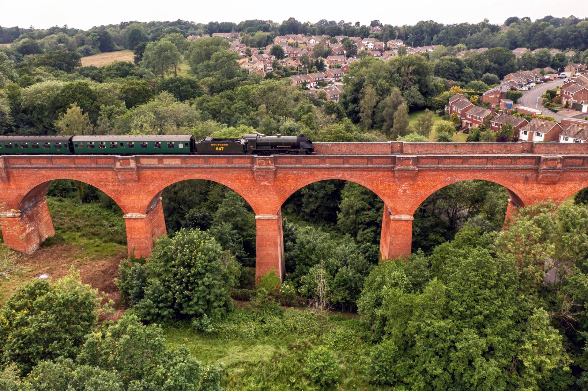 Aerial perspective of a vintage steam train crossing an iconic red brick arch viaduct surrounded by countryside hills, Love Locations TN22.