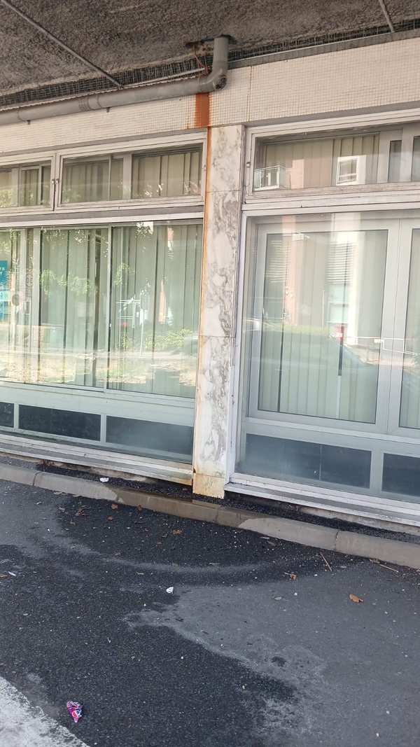 Exterior storefront and ground floor office film location in Watford WD18 available via Love Locations, featuring weathered modernist architecture, large glass display windows with vertical blinds, a marble-effect support pillar, and a concrete overhang.